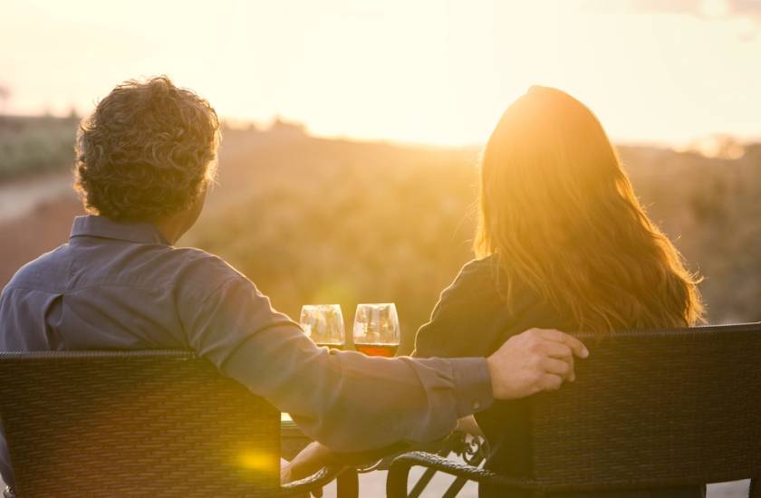 A couple sits at a winery with back toward the camera staring at sunset over vineyard